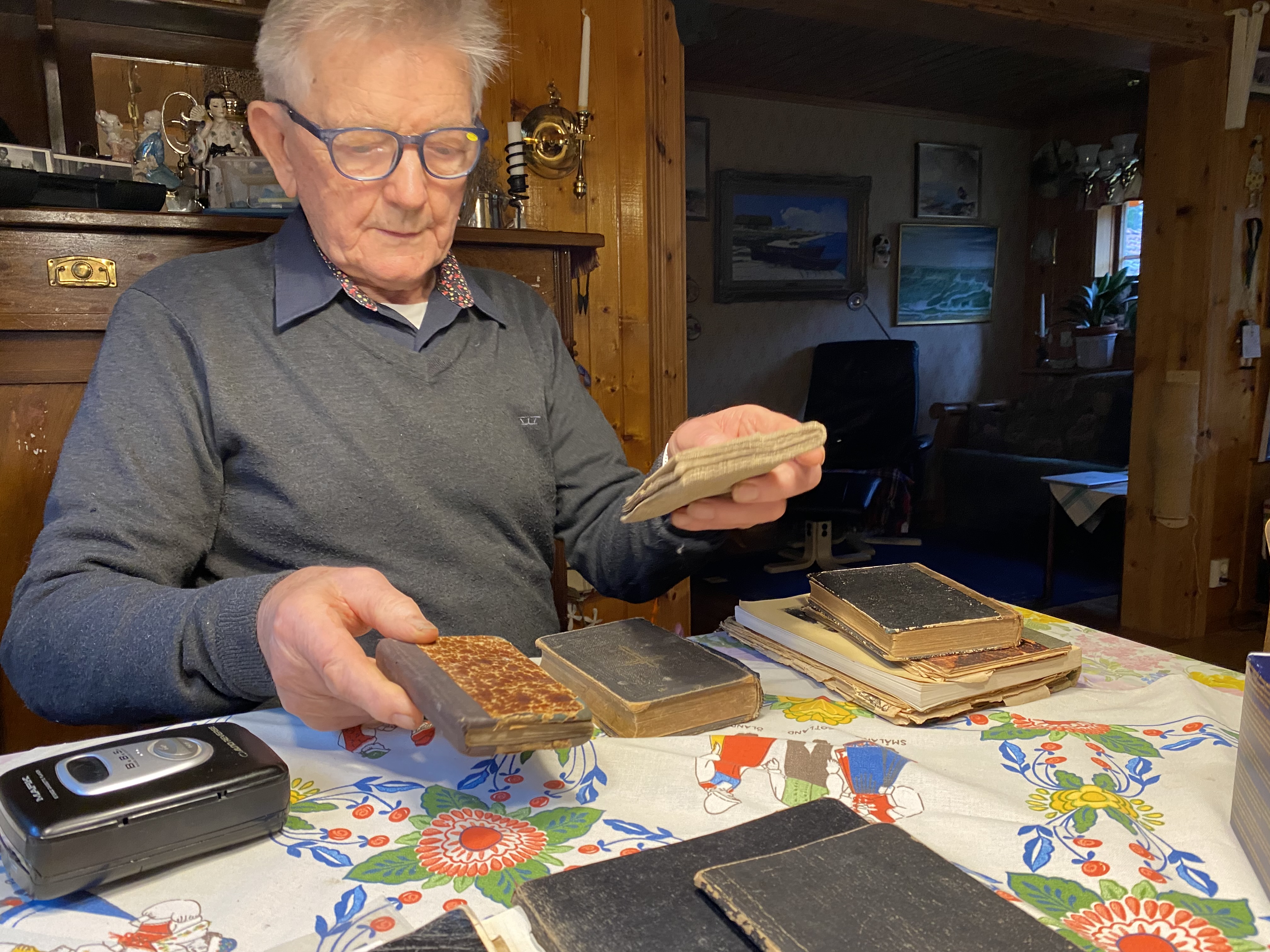 Fotografi av en man med glasögon som sitter vid ett bord och lyfter och ser på inbundna böcker. Bakom honom syns en stor byrå i trä och i rummet snett bakom hänger många tavlor.