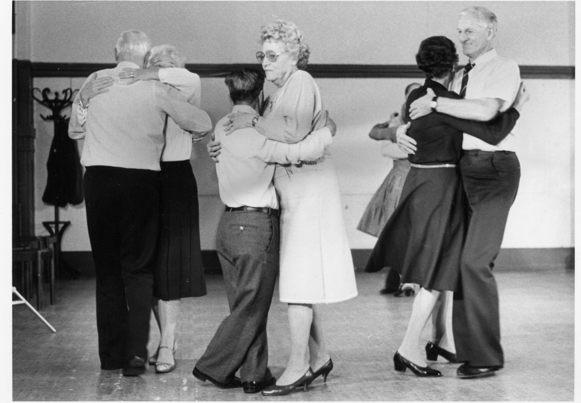 Svartvitt fotografi på tre äldre danspar.
    Paren dansar tätt ihop med båda händerna på parets rygg.