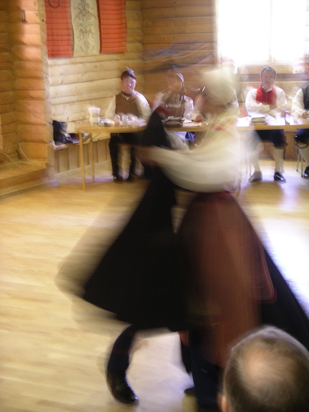 Fartfyllt och suddigt fotografi
    på dansande par i folkdräkt i förgrunden. Längre bak i den ljusa
    träbyggnaden sitter bedömningsnämnden vid ett bord, även de i
    folkdräkter.