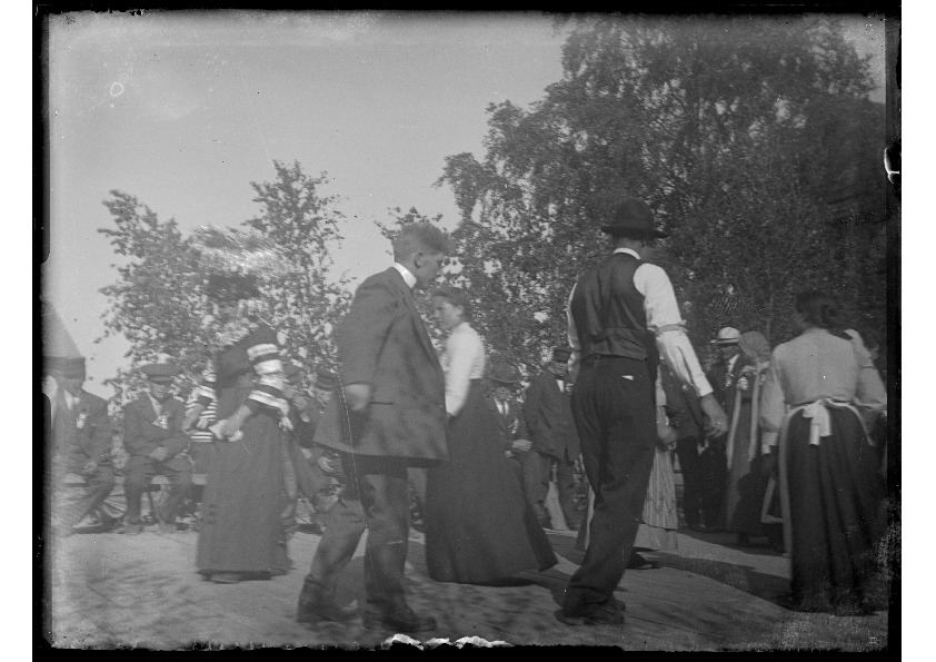 Svartvitt
    arkivfotografi av mänskor som dansar på ett trägolv under öppen himmel.
    Bilden är tagen vid midjehöjd, i bakgrunden syns lövträd och en lång
    bänkrad med åskådare.