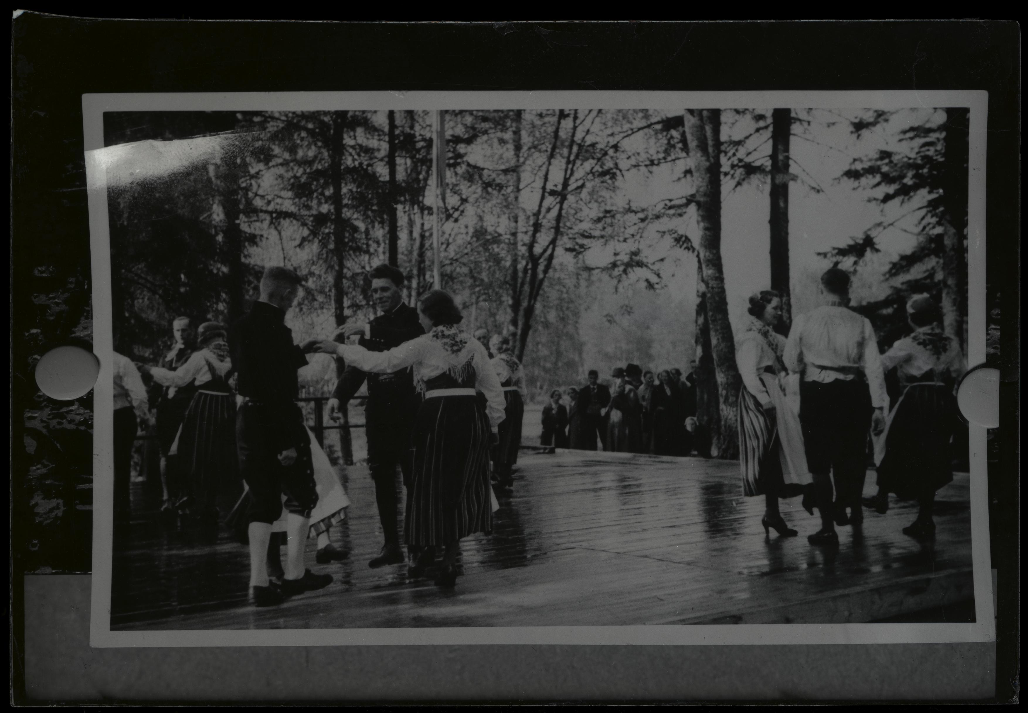 Mörkt,
    svartvitt arkivfotografi på tre dansande kvartetter utomhus. Längre bak,
    nedanför dansgolvet, står en klunga mänskor mellan trädstammar och ser
    på.